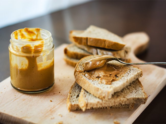 Pote de manteiga de amendoim e torradas barradas com manteiga de amendoim, empilhadas numa tábua de madeira.