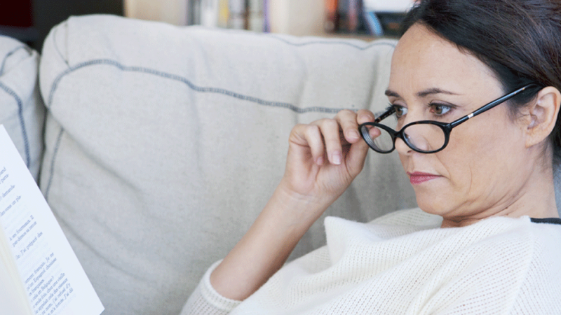 Mulher com vista cansada tenta ler livro no sofá