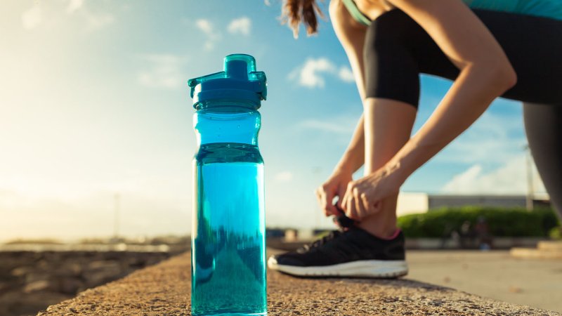 Garrafa de água em grande plano, com mulher a atar uma sapatilha no fundo.