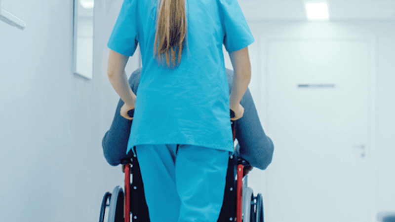 Profissional de saúde de uniforme azul, vista de costas, a empurrar uma pessoa numa cadeira de rodas por um corredor hospitalar branco e iluminado.
