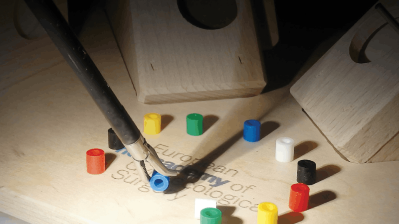 Base de madeira com pequenos cilindros coloridos espalhados, enquanto uma pinça segura uma peça azul. Ao fundo, há estruturas de madeira com buracos.