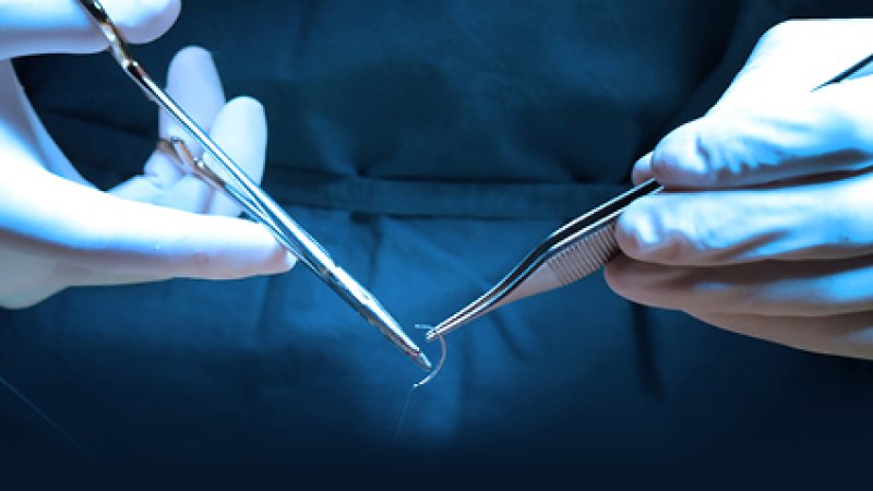Duas mãos com luvas cirúrgicas manuseando pinças médicas e uma agulha de sutura curvada sob iluminação azulada de centro cirúrgico.