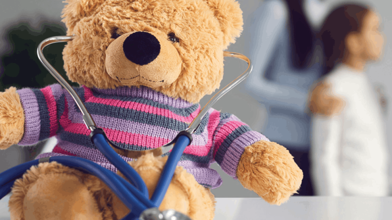 Um urso de peluche com uma camisola às riscas está sentado numa mesa branca com um estetoscópio azul à volta do pescoço. Ao fundo, uma mulher e uma criança aparecem desfocadas num consultório médico.