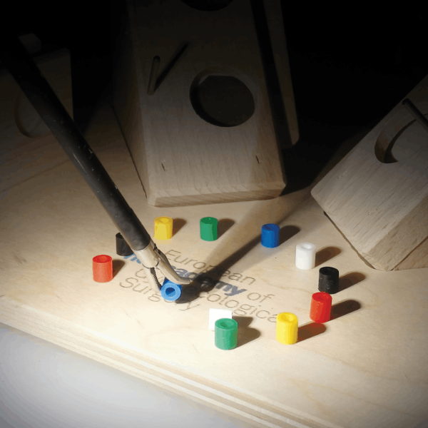 Base de madeira com pequenos cilindros coloridos espalhados, enquanto uma pinça segura uma peça azul. Ao fundo, há estruturas de madeira com buracos.