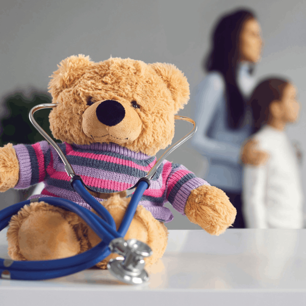Um urso de peluche com uma camisola às riscas está sentado numa mesa branca com um estetoscópio azul à volta do pescoço. Ao fundo, uma mulher e uma criança aparecem desfocadas num consultório médico.